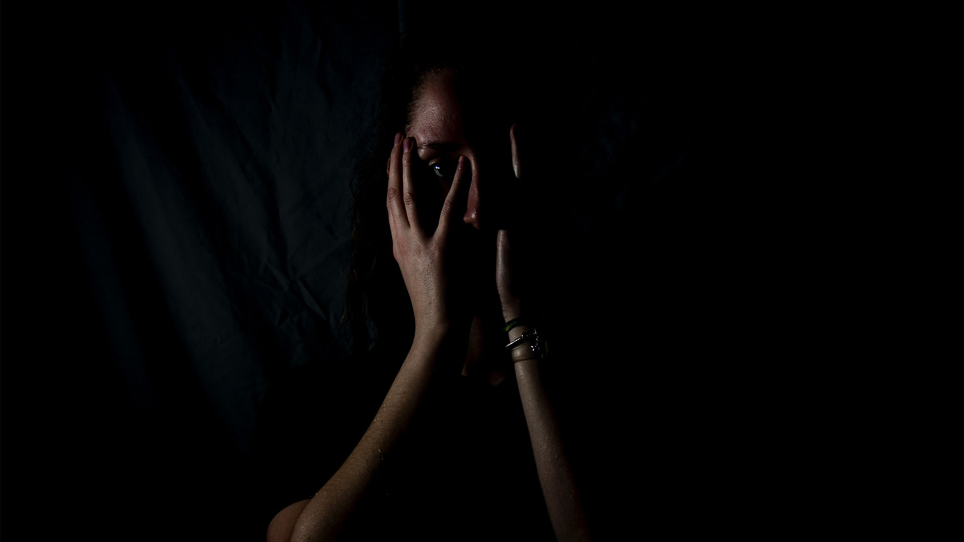 A woman holding her face with her hands in a dark background.